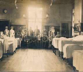 Patients in beds either side of ward and seated at the end with nurses