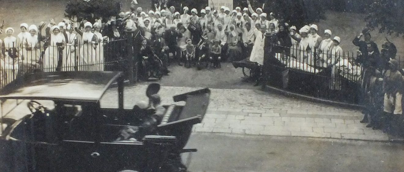 Old motor car driving past hospital staff and patients standing outside Infirmary gates waving