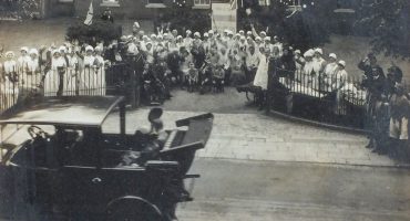 Old motor car driving past hospital staff and patients standing outside Infirmary gates waving