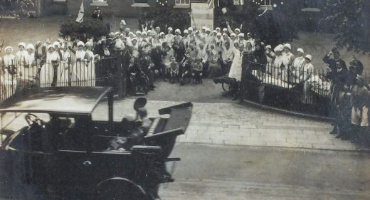 Old motor car driving past hospital staff and patients standing outside Infirmary gates waving