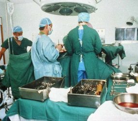 Surgeons and anaethetist in greens, operating on patient, equipment in foreground, pelvis X-ray on viewers in background