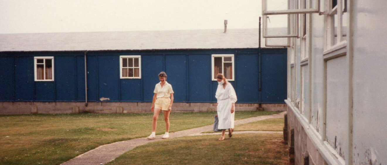 1 person in shorts and scientist in white gown and mask walk on grass next to hut