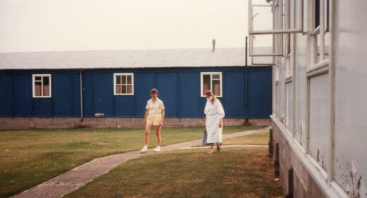 1 person in shorts and scientist in white gown and mask walk on grass next to hut