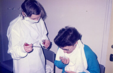 Colour photograph showing scientist in gown and mask next to seated person