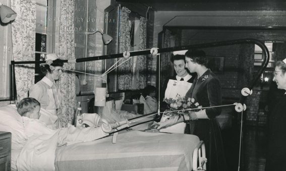 Princess with nurses talking to boy in traction on a bed in ward