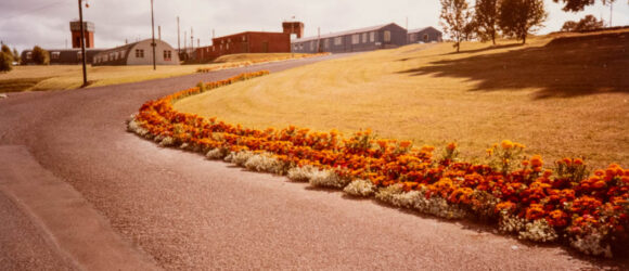 A colour photograph of curved drive, flower bed and huts in distance