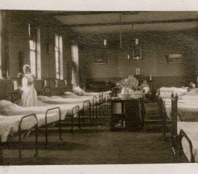Nurse doing her rounds, metal beds line both sides of the ward