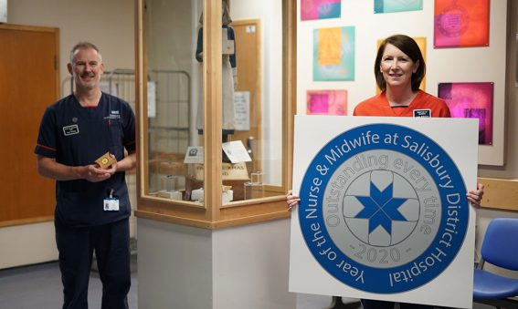 Lorna holding large badge design print out, standing next to display cabinet with Henry