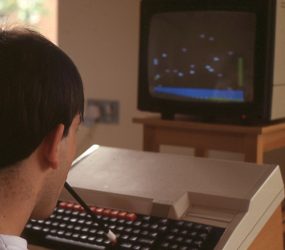 seated man with rubber ended pencil in mouth tapping on large keyboard attached to large TV screen monitor showing simple graphics