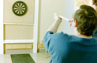 man in wheelchair blowing a pipe with dart aimed at dart board