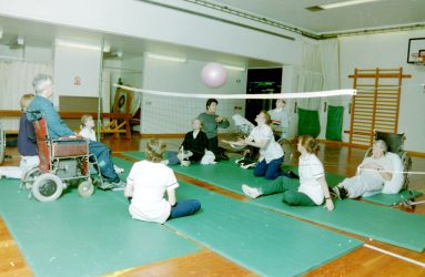 physiotherapists and occupational therapists sit on floor with patients some in wheelchairs, some on the floor playing volleyball in gym
