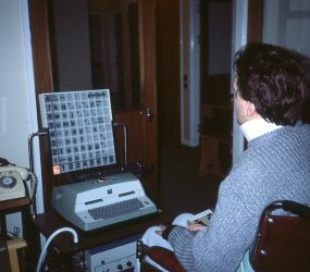 man in wheelchair in front of large typewriter device with a light box above it displaying letters and numbers in a grid